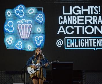 A woman singing with a guitar. Behind her is a neon display saying Lights! Canberra! Action! @ Enlighten Festival